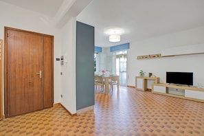 A spacious living area with a wooden door, a dining table with chairs, a flat-screen TV, and a patterned floor.