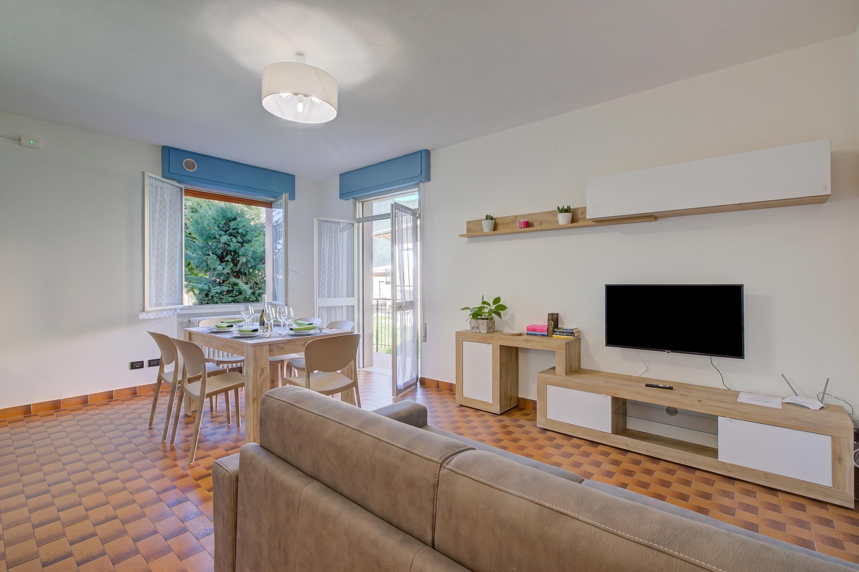A modern living room with a sofa, a TV, a dining area with chairs and a table, and a balcony with a view of greenery.