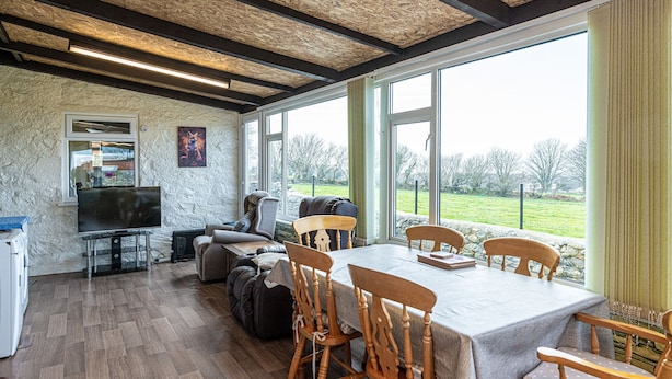 A dining area with a table set for six, wooden chairs, and a view of a green landscape through large windows. - Fox's Lair Farmhouse - sleeps 6 guests  in 3 bedrooms (Caernarfon)