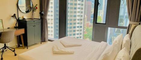A hotel room with a large bed, a desk with a chair, a round mirror, and a view of tall buildings through the window.