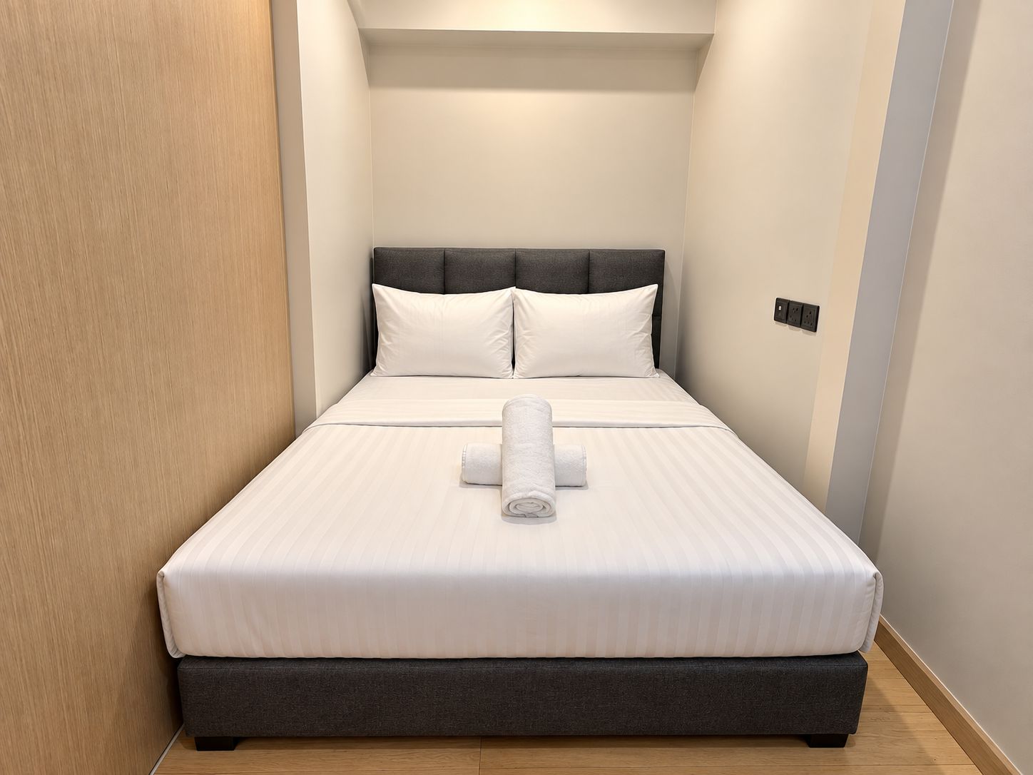 A neatly made bed with white linens and a rolled towel, a dark headboard, and a light-colored wall with a switch.