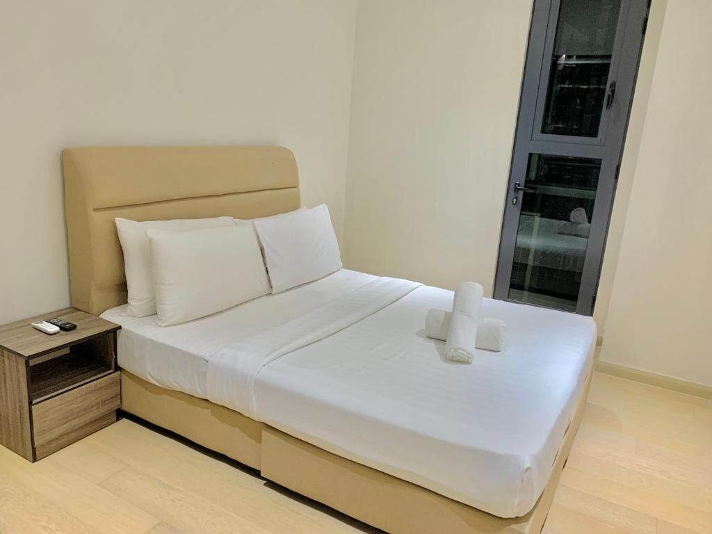 Una cama bien hecha con sábanas y almohadas blancas, una mesita de noche de madera con un control remoto, y una puerta de vidrio a la derecha.