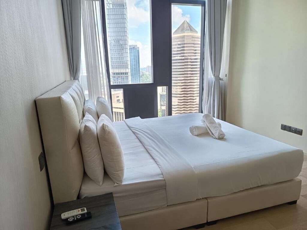 A hotel room with a large bed, a remote control on the nightstand, and a view of tall buildings through the window.