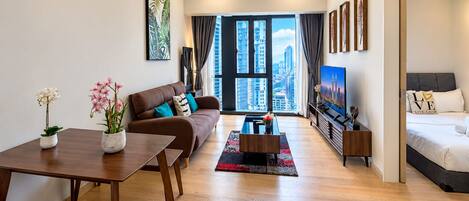 A modern living room with a sofa, coffee table, and a TV. There is a dining table with chairs, a large window with a city view, and a bed with a cityscape poster.