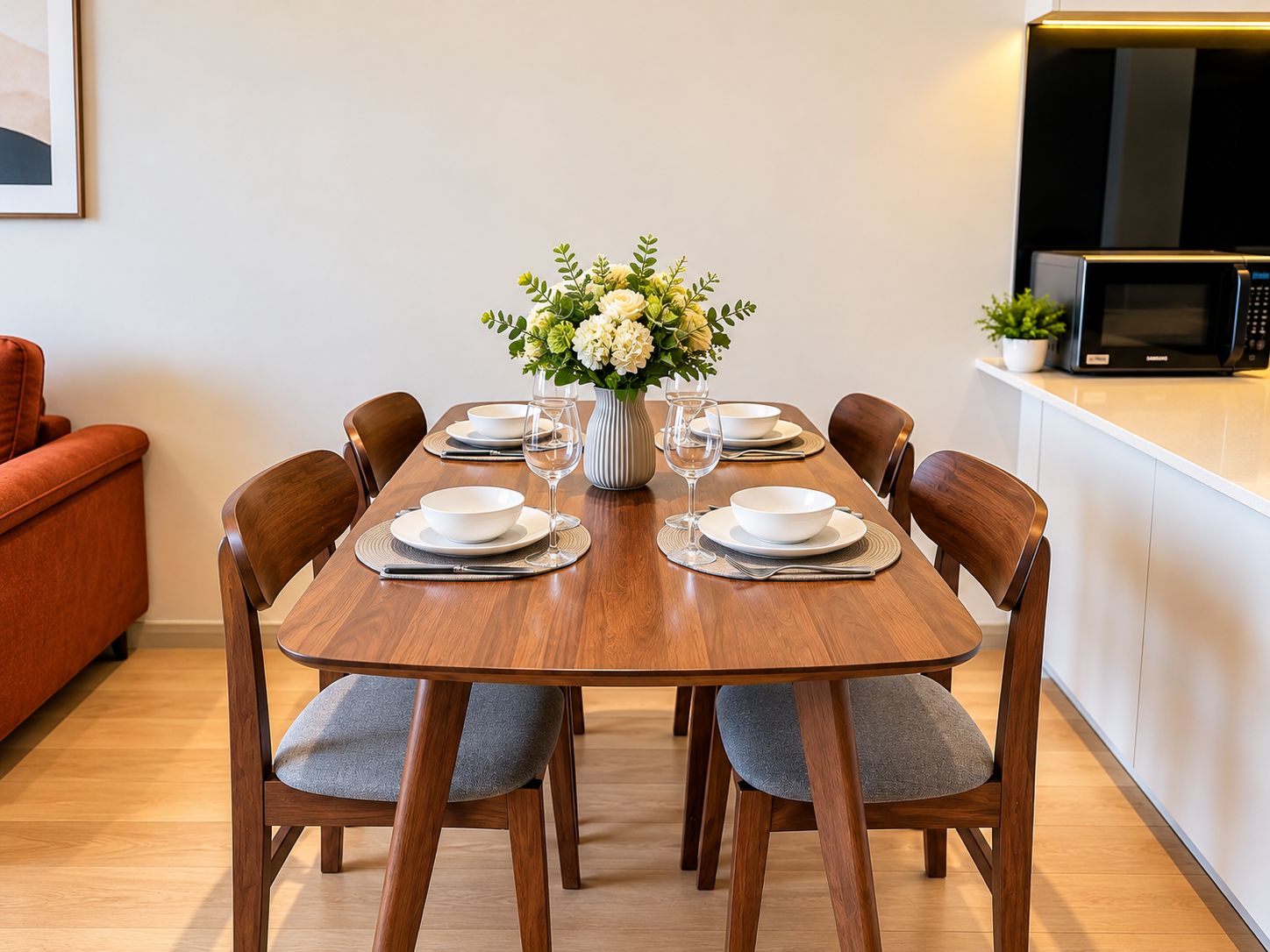 A dining table set for four with a vase of flowers, a microwave, and a sofa in the background.