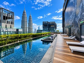 Una piscina en la azotea con sillones de descanso, vistas a las Torres Petronas y un edificio moderno con fachada de vidrio.