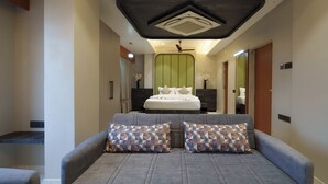 A modern bedroom with a grey sofa, patterned pillows, a bed with white bedding, and a ceiling fan. - Hotel Hari Om (Dwarka)
