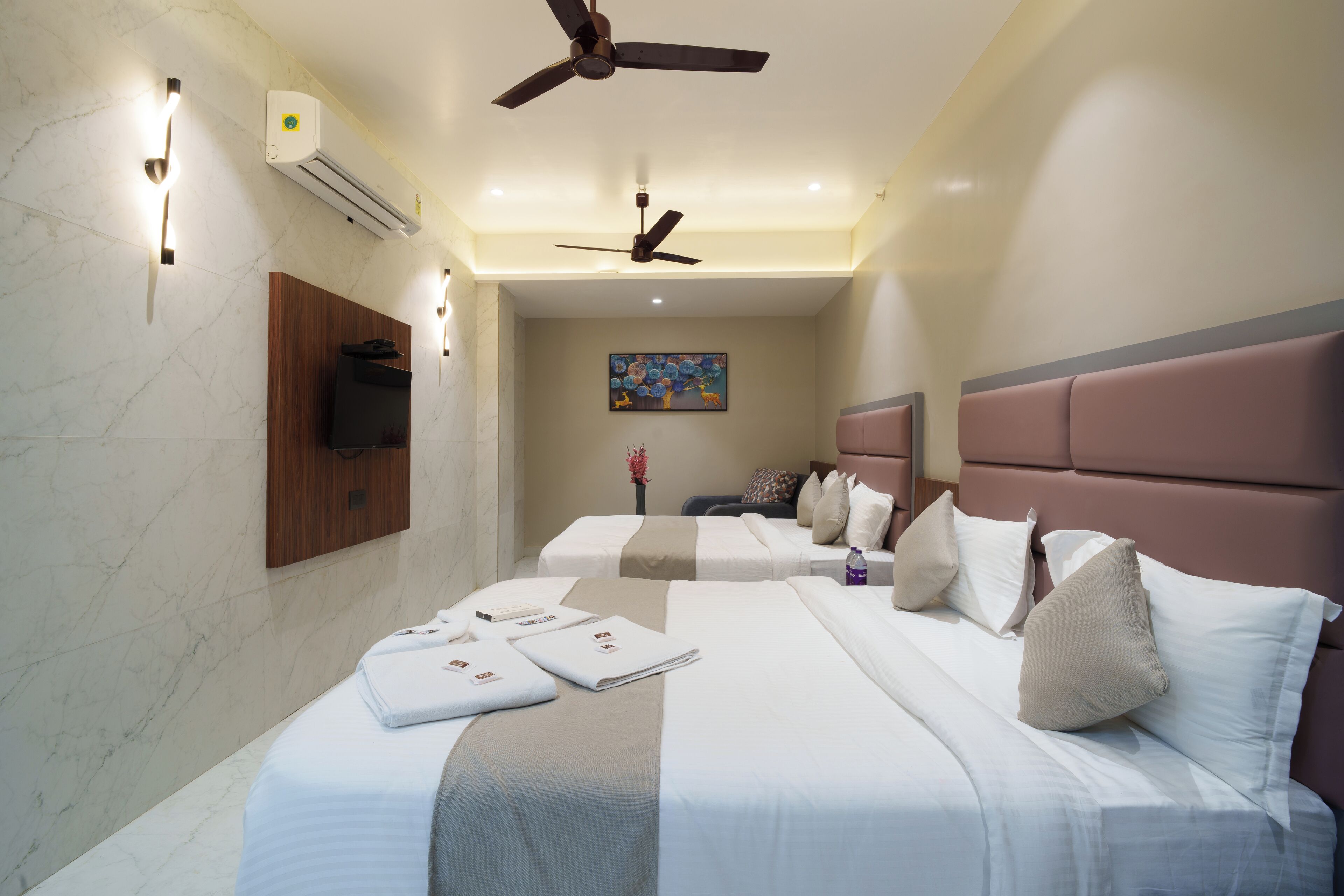 Habitación de hotel con dos camas, ventilador de techo, aire acondicionado, televisor montado y un cuadro en la pared.