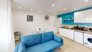 Un espacio habitable compacto con kitchenette, un sofá azul, una zona de comedor y un reloj de pared.