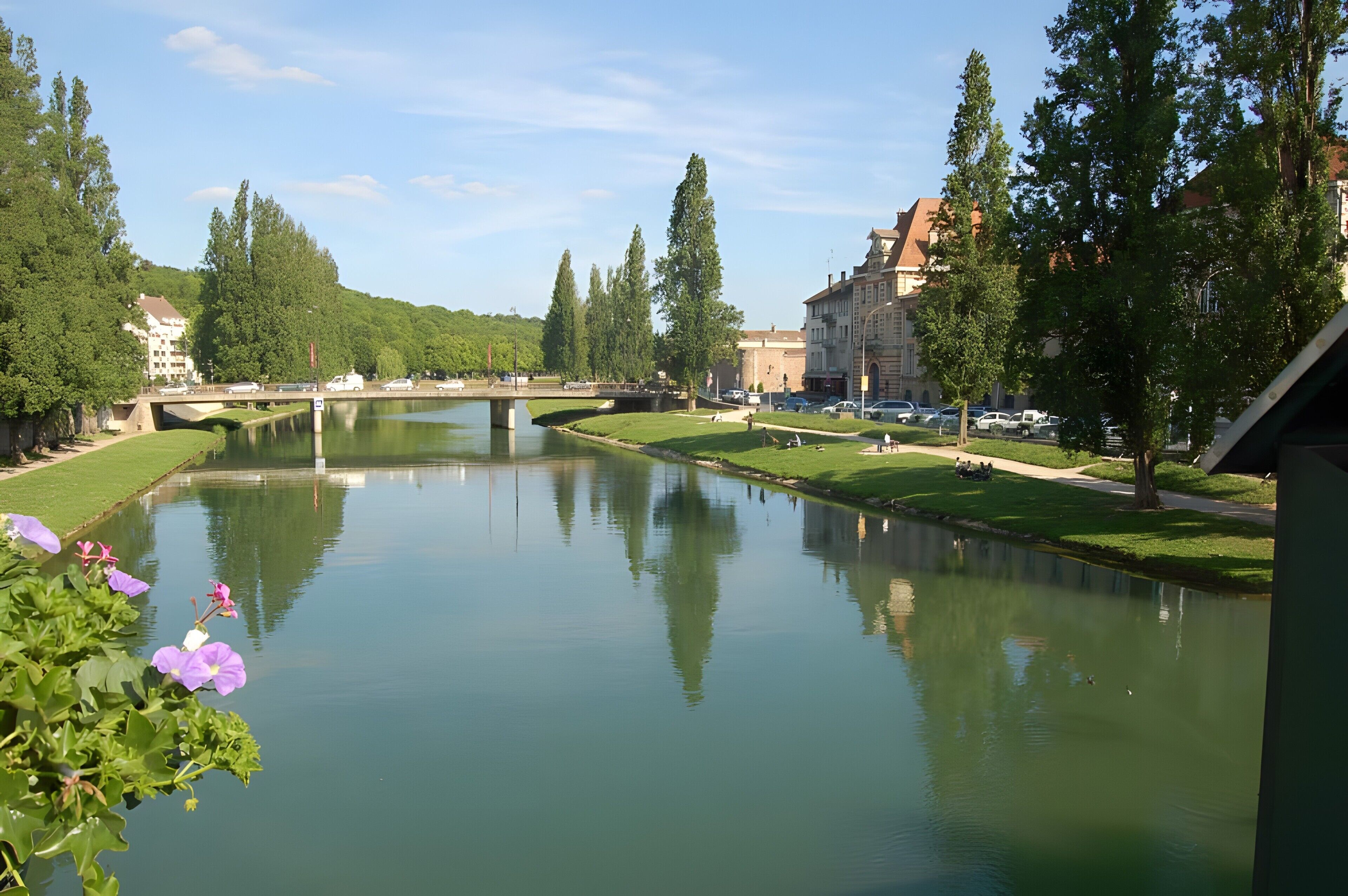 A calm river with a bridge, greenery, and buildings in the background.