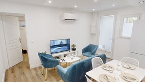A modern living room with a TV, two blue chairs, a dining table set for four, and a white air conditioning unit. - Apartment 'Cosy Calm Seine Riverside Garden' with Balcony and Wi-Fi (Le Mée-sur-Seine)