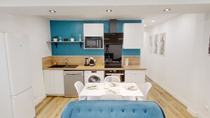 A compact kitchen with a microwave, sink, and dishwasher, a dining area with a table set for two, and a blue upholstered bench. - Apartment 'Cosy Calm Seine Riverside Garden' with Balcony and Wi-Fi (Le Mée-sur-Seine)