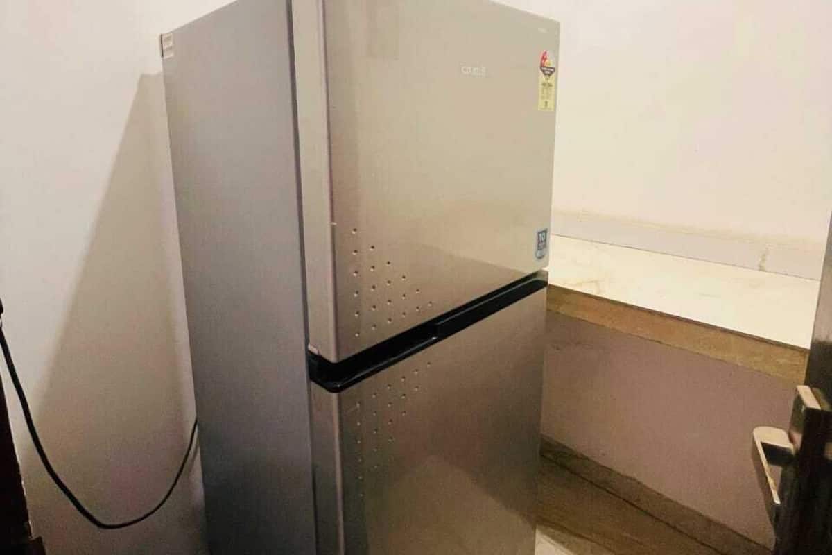 Un refrigerador compacto de acero inoxidable en un rincón de una habitación con pared blanca y piso de madera.