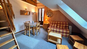 Een gezellig zolderkamertje met een houten trap, een eettafel met stoelen, een kleine bank en een lamp.