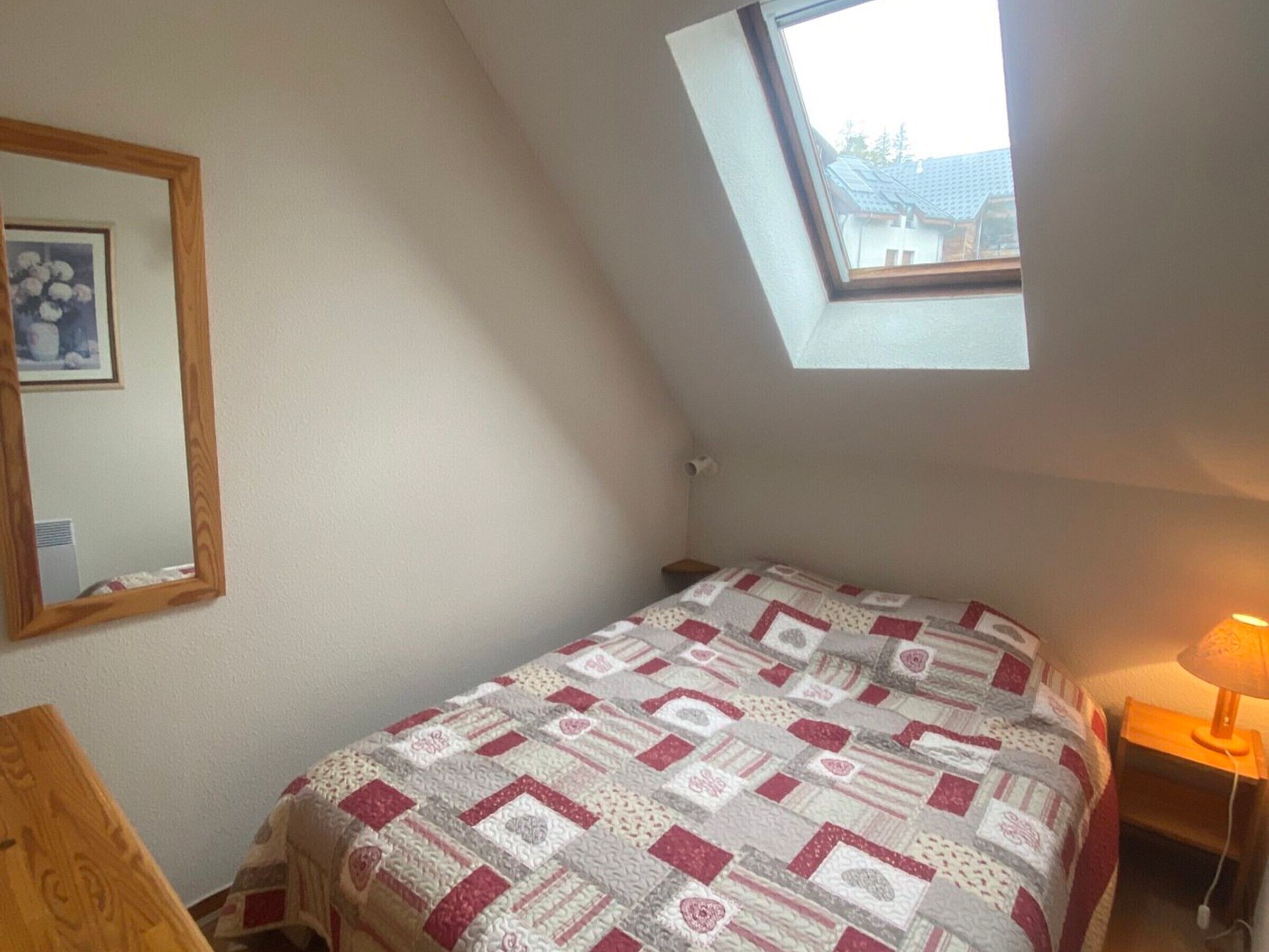 A bedroom with a sloped ceiling, a skylight, a bed with a patterned quilt, a bedside table with a lamp, and a mirror on the wall.