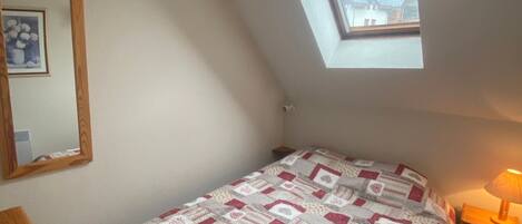A bedroom with a sloped ceiling, a skylight, a bed with a patterned quilt, a bedside table with a lamp, and a mirror on the wall.