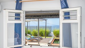 A room with an open window revealing a pool area with lounge chairs and a pergola.