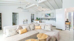 A modern living room with a sectional sofa, a round wooden coffee table, and a kitchen area with white cabinets and a tiled backsplash.