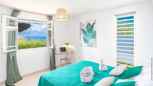 A bedroom with a view of the sea, a desk with a laptop, a bed with green bedding, and a hanging light fixture.