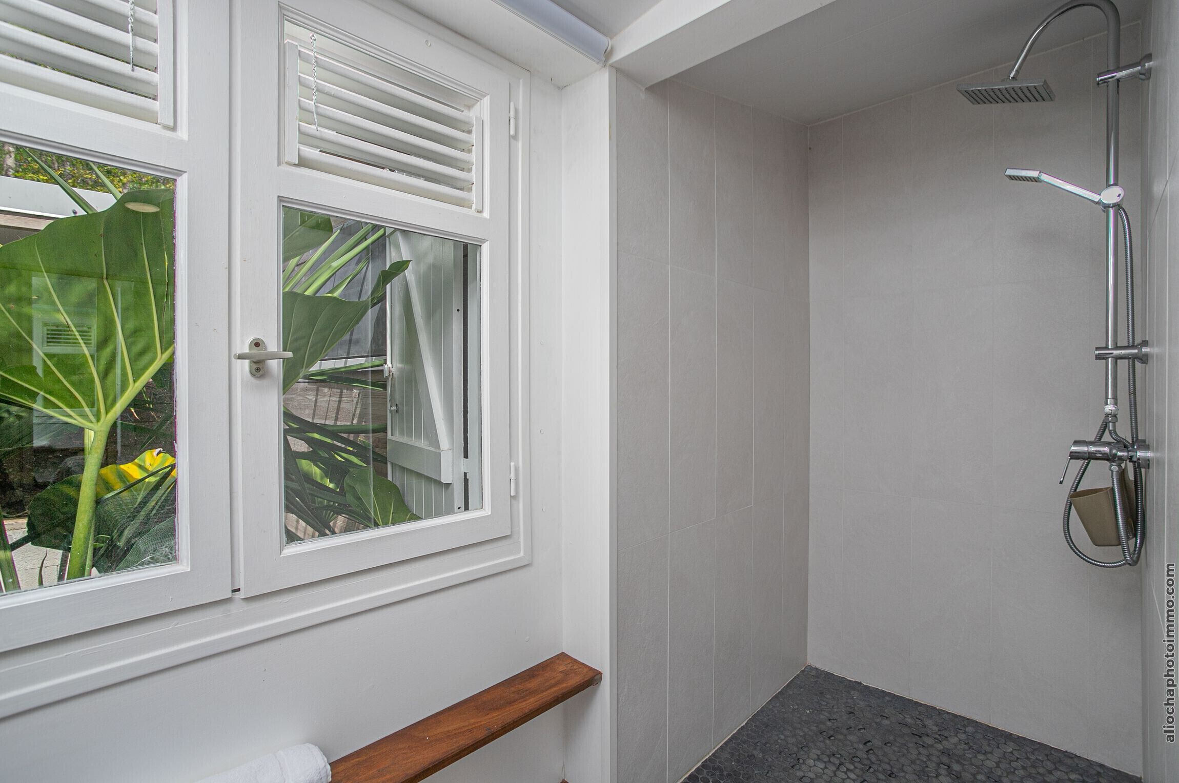 A bathroom with a shower, a window with blinds, and a wooden shelf.