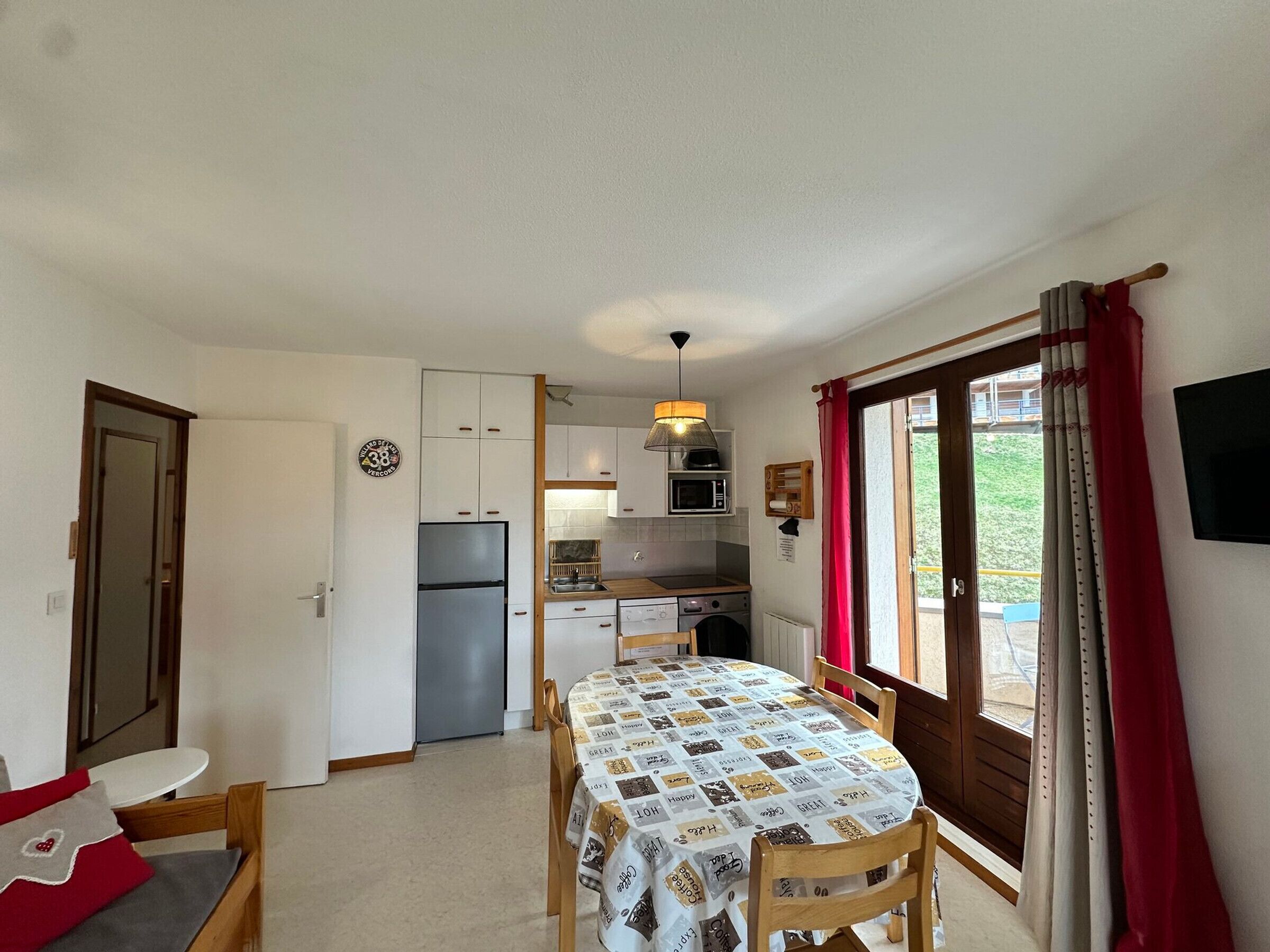 A compact kitchen with a refrigerator, microwave, and sink. A dining table with chairs is positioned near a window with curtains.