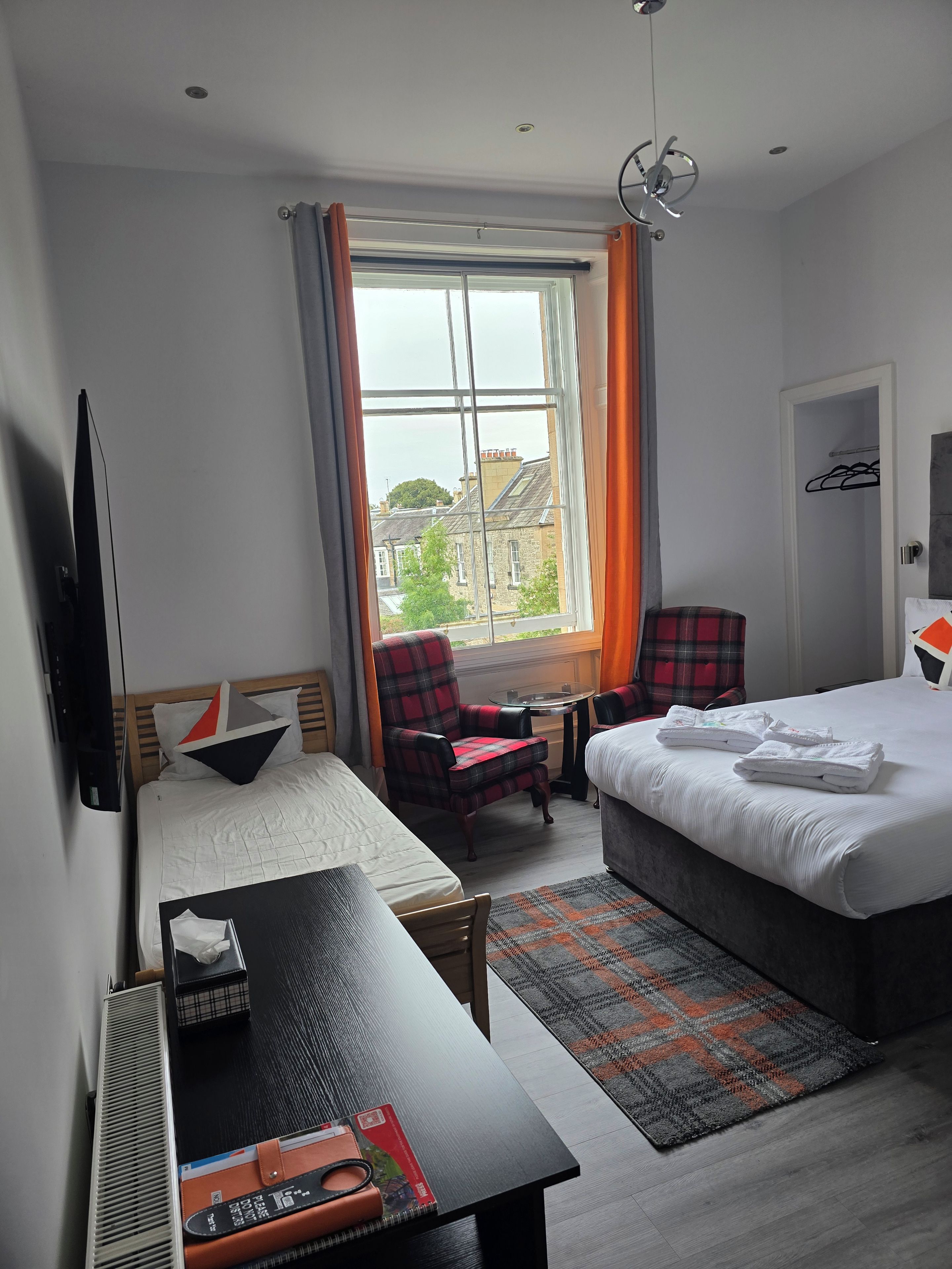 A hotel room with a bed, a desk with a chair, a TV, a window with curtains, and a view of buildings outside.