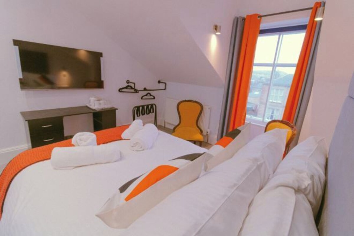 Une chambre d’hôtel avec un lit, un téléviseur fixé au mur, une chaise, un bureau et une fenêtre avec des rideaux.