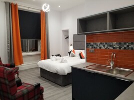 A hotel room with a bed, a small kitchenette, a chair, and a window with curtains.