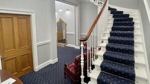 A grand staircase with a wooden handrail and blue carpeted steps, leading to a landing with a chandelier and a mirror. - Mayfield Suites  (Edinburgh)
