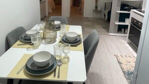 A dining area with a table set for four, chairs, and a kitchen area with shelves and appliances. - Portu Sereno Emerald - Floor 1 (Setúbal)