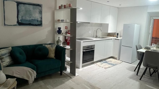 A modern living room with a green sofa, a dining area with a table and chairs, and a kitchen with white cabinets and appliances. - Portu Sereno Emerald - Floor 1 (Setúbal)