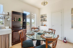 A dining area with a glass table, wooden chairs, and a kitchen area with shelves and utensils. - Villa Laura by Rentbeat  (Ostuni)