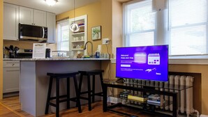 A modern kitchen with a central island, bar stools, a flat-screen TV, and a dining area with a window providing natural light. - Comfy 1 BR Hide Away Apt w/ Garage Parking (Bryn Mawr)