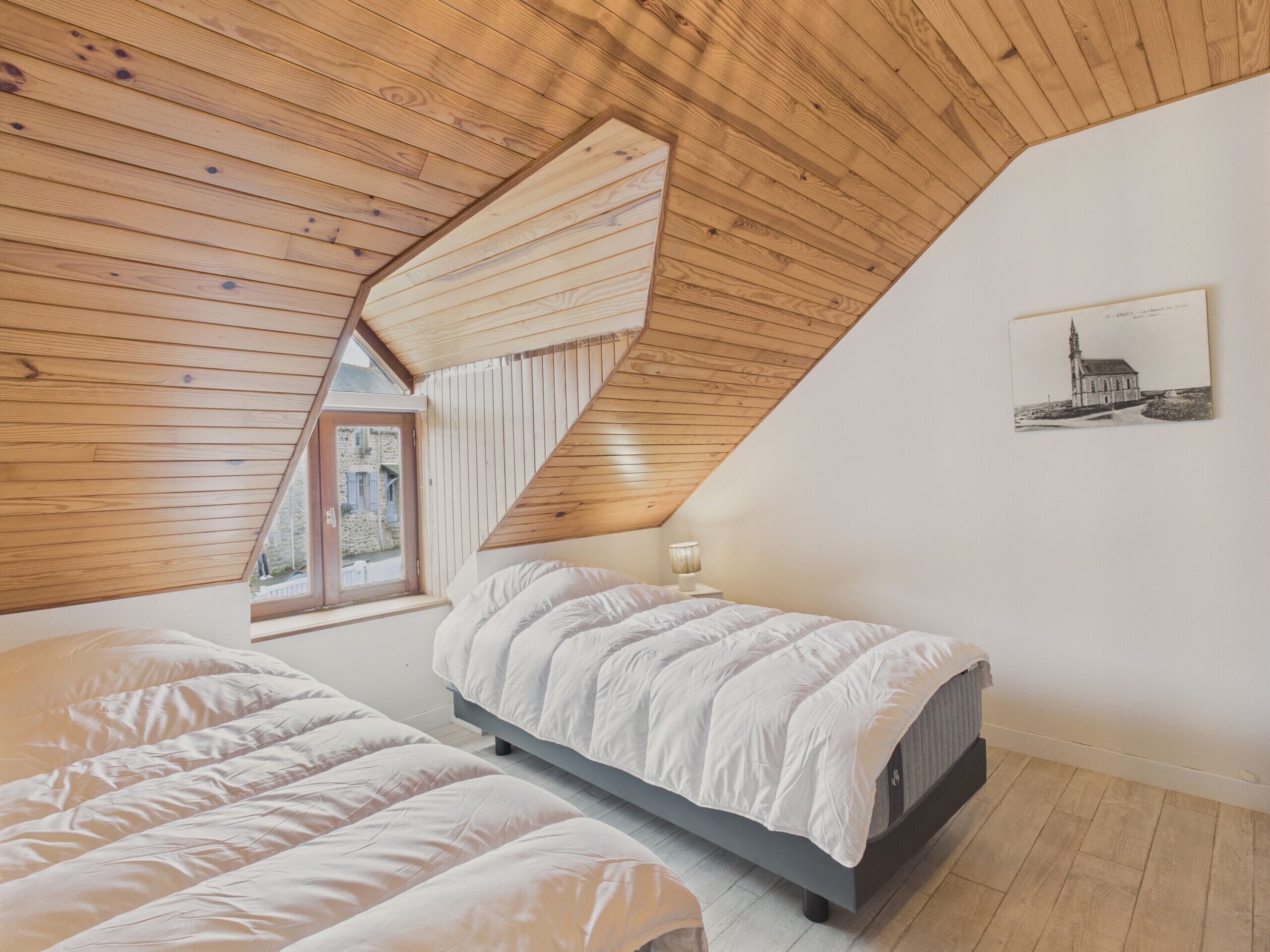 Une chambre avec deux lits, un plafond en pente avec des poutres apparentes, une fenêtre et un tableau accroché au mur.
