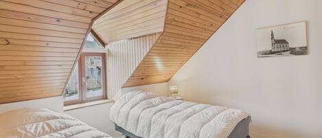 Une chambre avec deux lits, un plafond en pente avec des poutres apparentes, une fenêtre et un tableau accroché au mur.