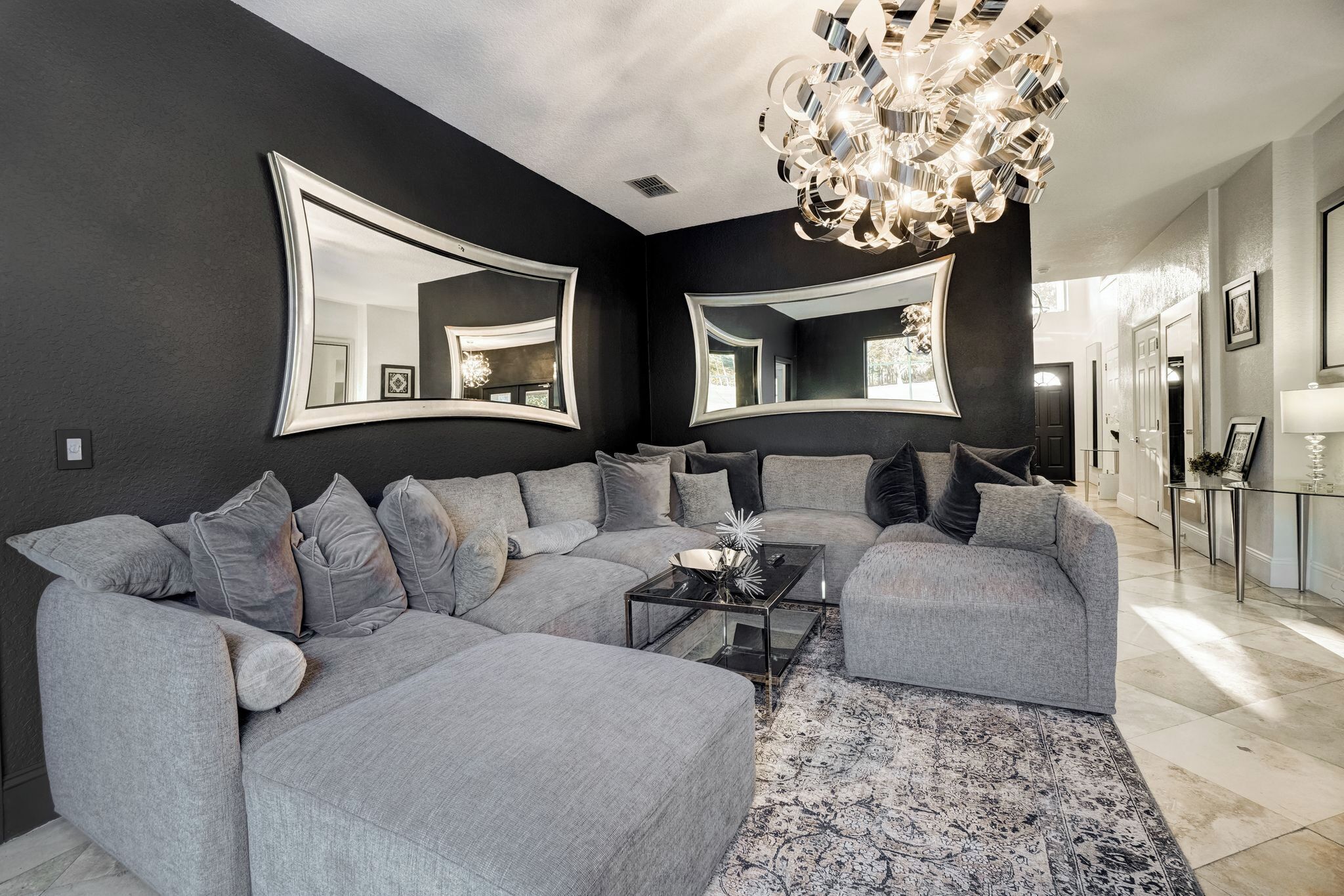 A modern living room with a grey sectional sofa, a glass coffee table, and a patterned rug. There are two large mirrors on the wall and a chandelier.