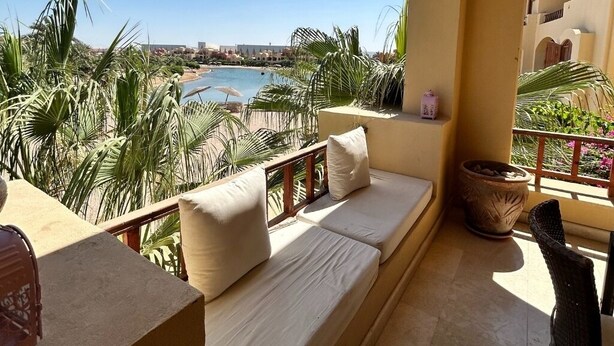 A balcony with a sofa, a chair, and a view of a beach and palm trees.