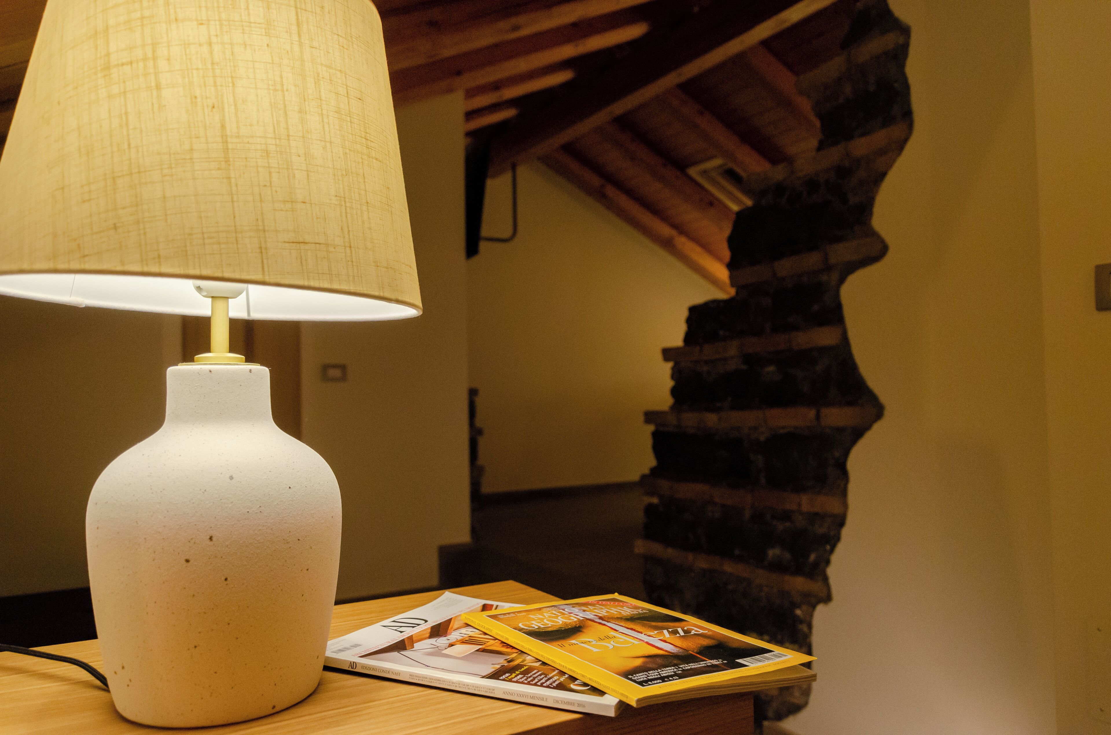 Une lampe de table blanche avec un abat-jour beige, un magazine posé sur la table et un escalier en bois en arrière-plan.