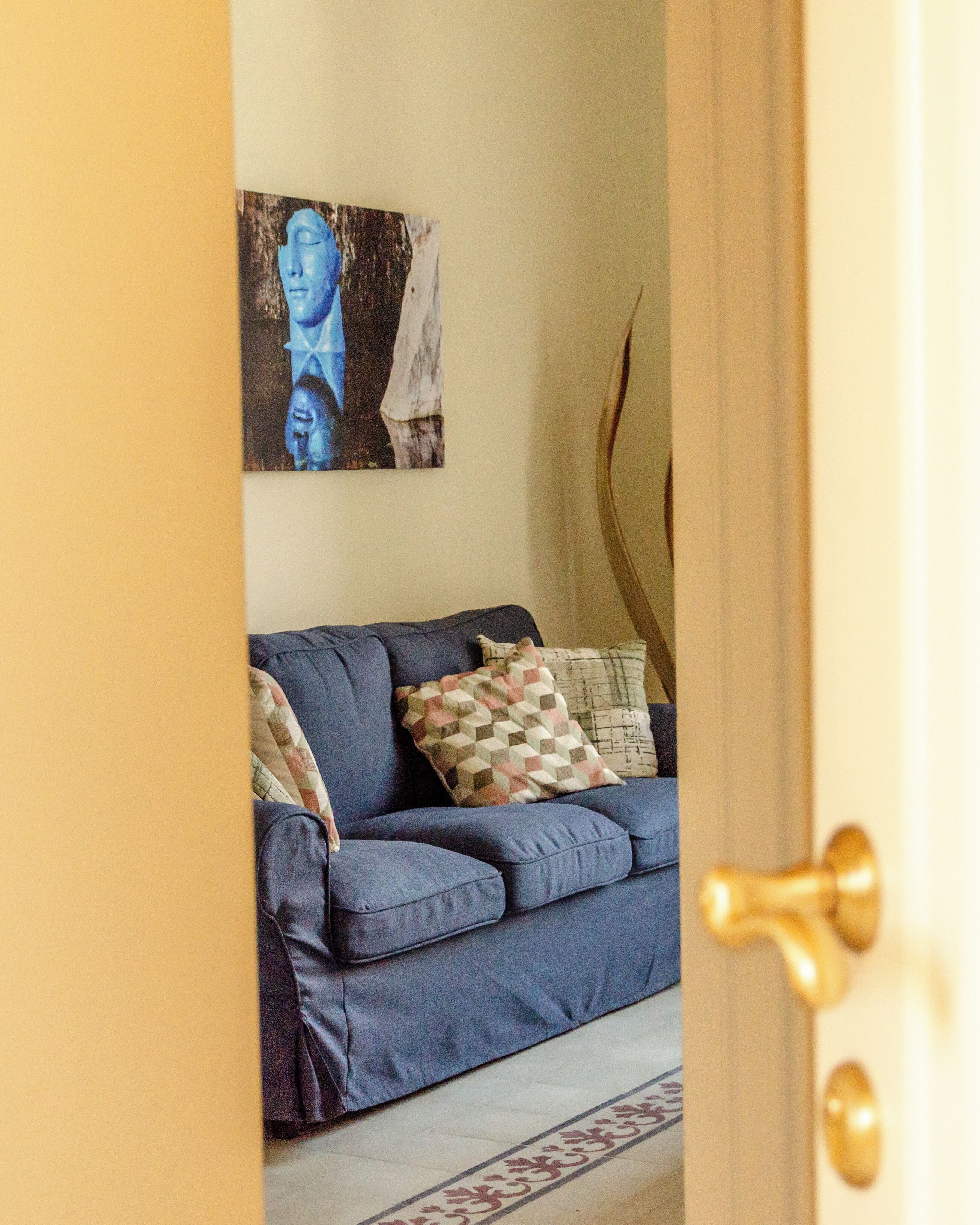 Une pièce avec un canapé bleu, des coussins à motifs, un tableau accroché au mur et une porte dotée d’une poignée en laiton.