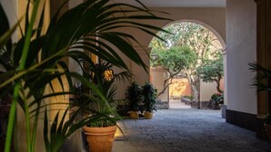 Une cour dallée de pierres, agrémentée de plantes en pot et surmontée d’une arche menant à un jardin.