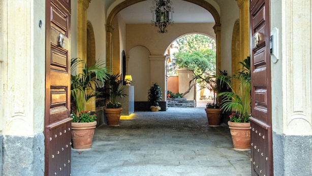 Un couloir dallé de pierres, avec des portes en arc de cercle, des plantes en pot et un luminaire suspendu.