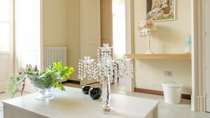 Une pièce avec une table blanche, un vase en verre contenant des fleurs, un radiateur blanc, un seau blanc et un tableau encadré accroché au mur.