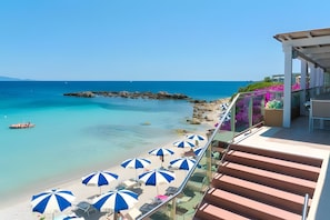 A balcony with a view of a beach, clear blue water, and a rocky outcrop.