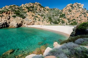 A coastal landscape with a clear turquoise sea, white sandy beach, and rocky cliffs.