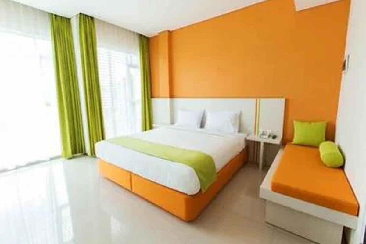 A hotel room with a bed, a bedside table, and a seating area with a cushion.