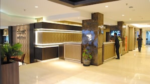 A modern hotel reception area with a marble floor, a reception desk, a large potted plant, and a television screen. - La Asiana Express Hotel (Bandung)