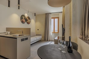 A modern hotel room with a kitchenette, a sofa, a dining table with wine glasses and a bottle, and a large window with curtains.