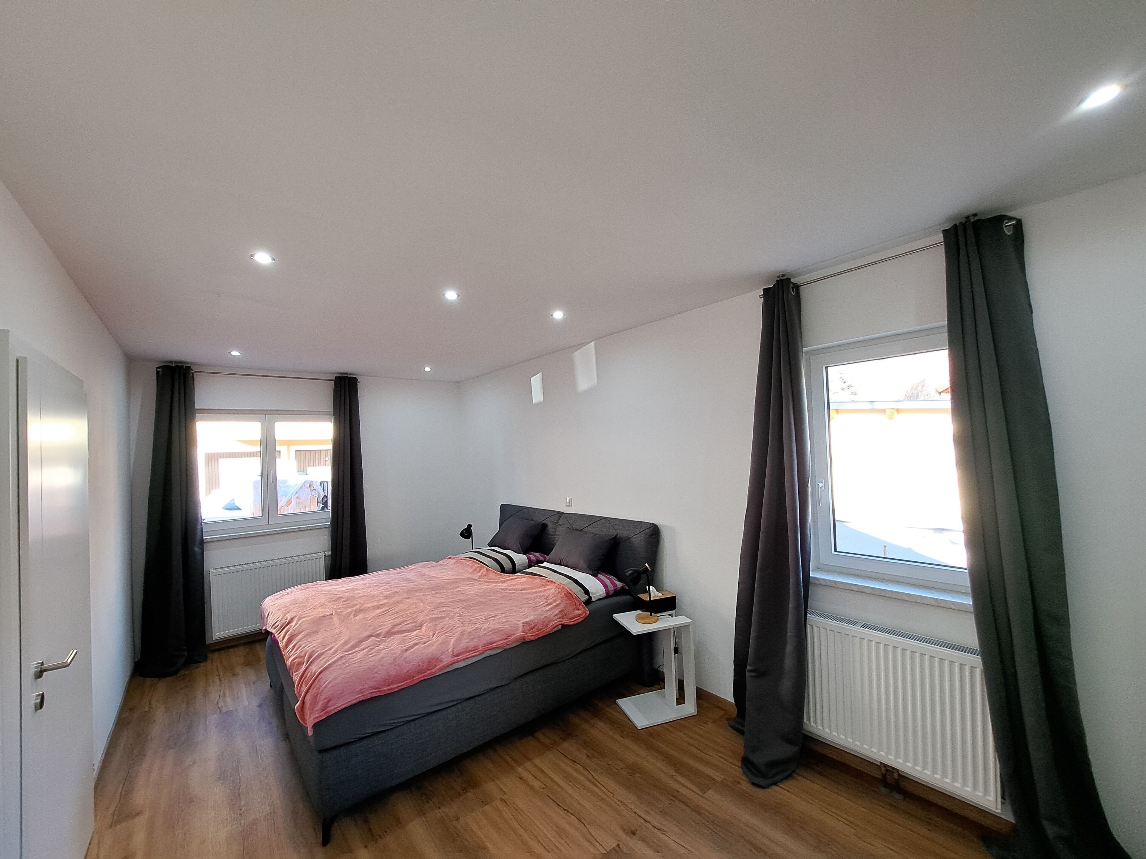 Un dormitorio con cama, mesita de noche, ventana con cortinas, radiador y puerta.