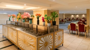 Un vestíbulo de hotel con mostrador de recepción, plantas en macetas y un área de comedor con mesas y sillas.
