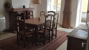 A dining area with a wooden table and chairs, a red patterned rug, a fireplace, a lamp, and a tall wooden clock. - Le Balcon d'Arcyate (Arcy-sur-Cure)